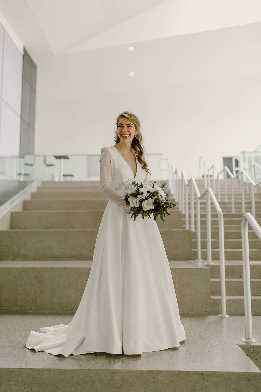 Women in long sleeve v-neck white wedding dress holding green and white flowers on concrete stairs